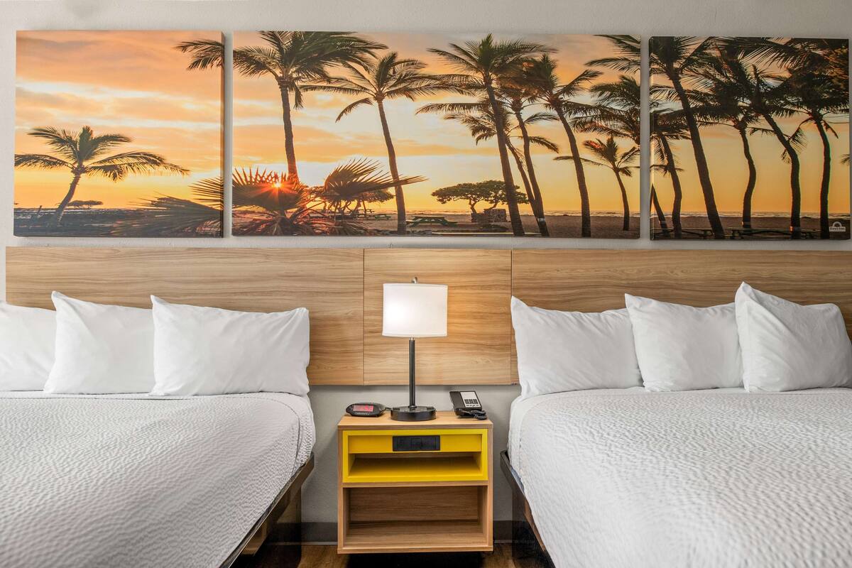 Une chambre d’hôtel avec deux lits, une tête de lit en bois, une table de chevet avec une lampe et un tableau représentant des palmiers au coucher du soleil, accroché au mur.