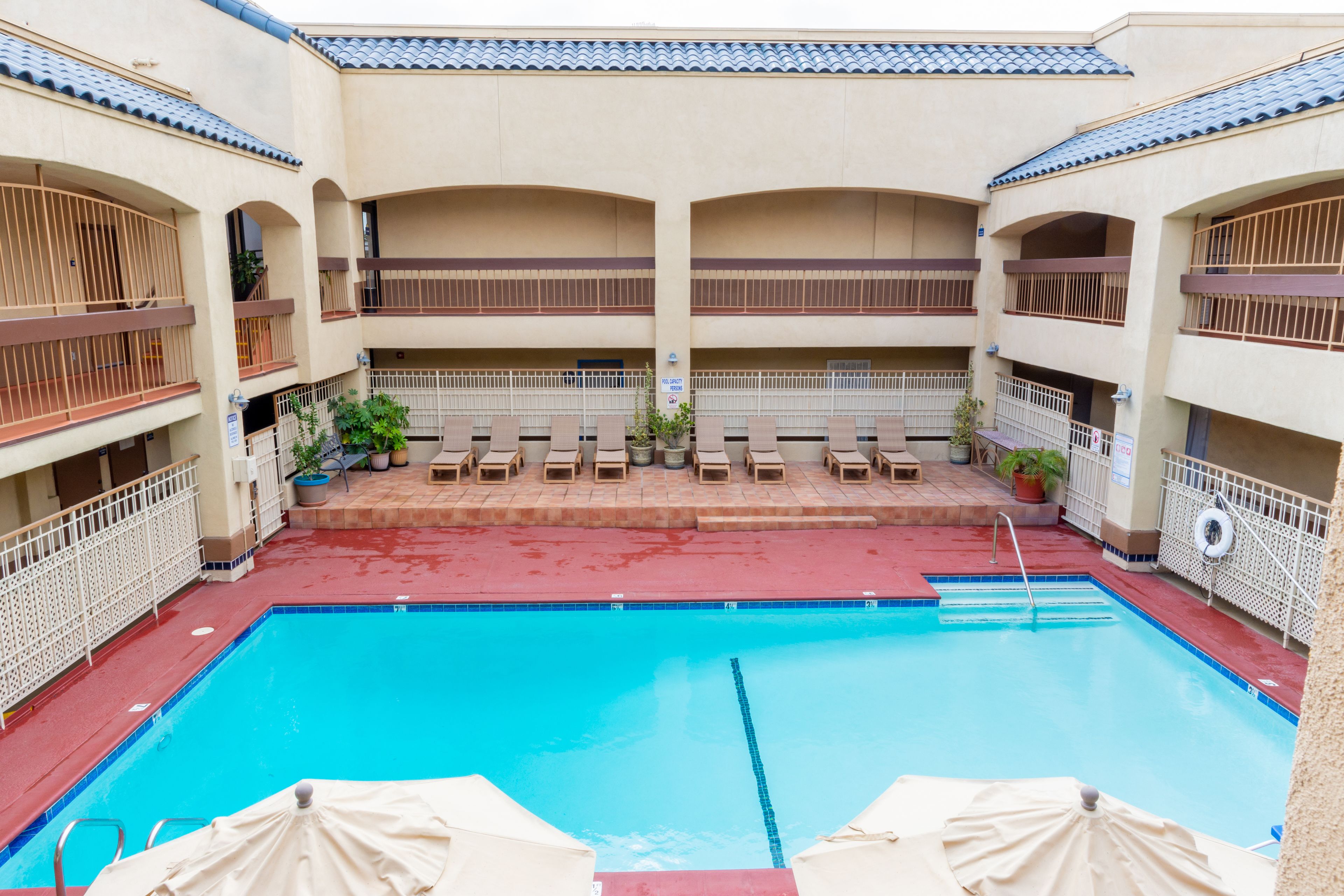A swimming pool with lounge chairs and umbrellas, surrounded by a building with multiple balconies and a tiled roof.