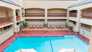 A swimming pool with lounge chairs and umbrellas, surrounded by a building with multiple balconies and a tiled roof.