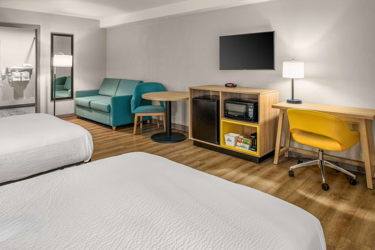 Une chambre d’hôtel avec deux lits, un canapé de couleur turquoise, une petite table ronde, un four à micro-ondes, une télévision et une chaise jaune.