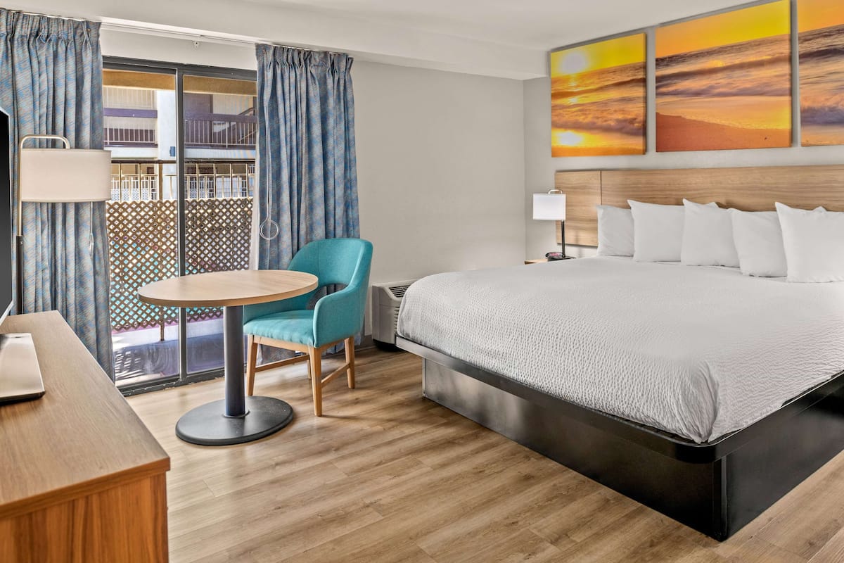 Une chambre d’hôtel avec un grand lit, une table ronde, une chaise bleue et une vue sur un balcon doté d’une balustrade.