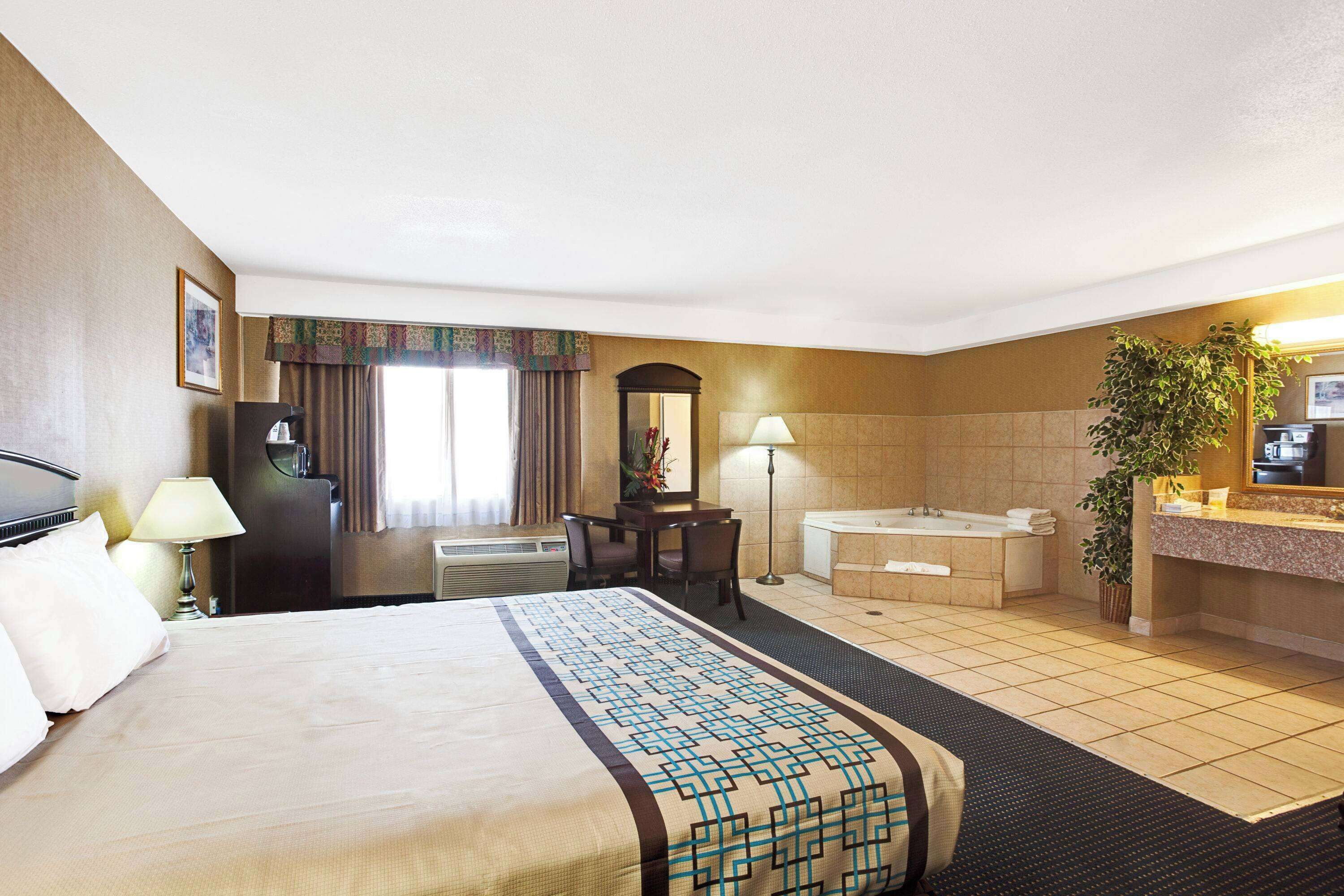 Une chambre d’hôtel comprenant un lit, un bureau, une chaise, une lampe de bureau, un miroir et une salle de bain avec une baignoire et un lavabo.