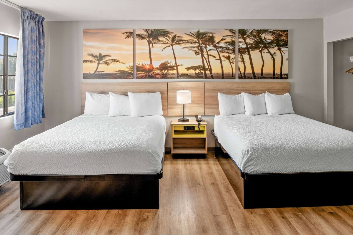 Une chambre d’hôtel avec deux lits, un plancher en bois, un mur orné d’un tableau repr ésentant des palmiers au coucher du soleil, et une table de chevet avec une lampe.