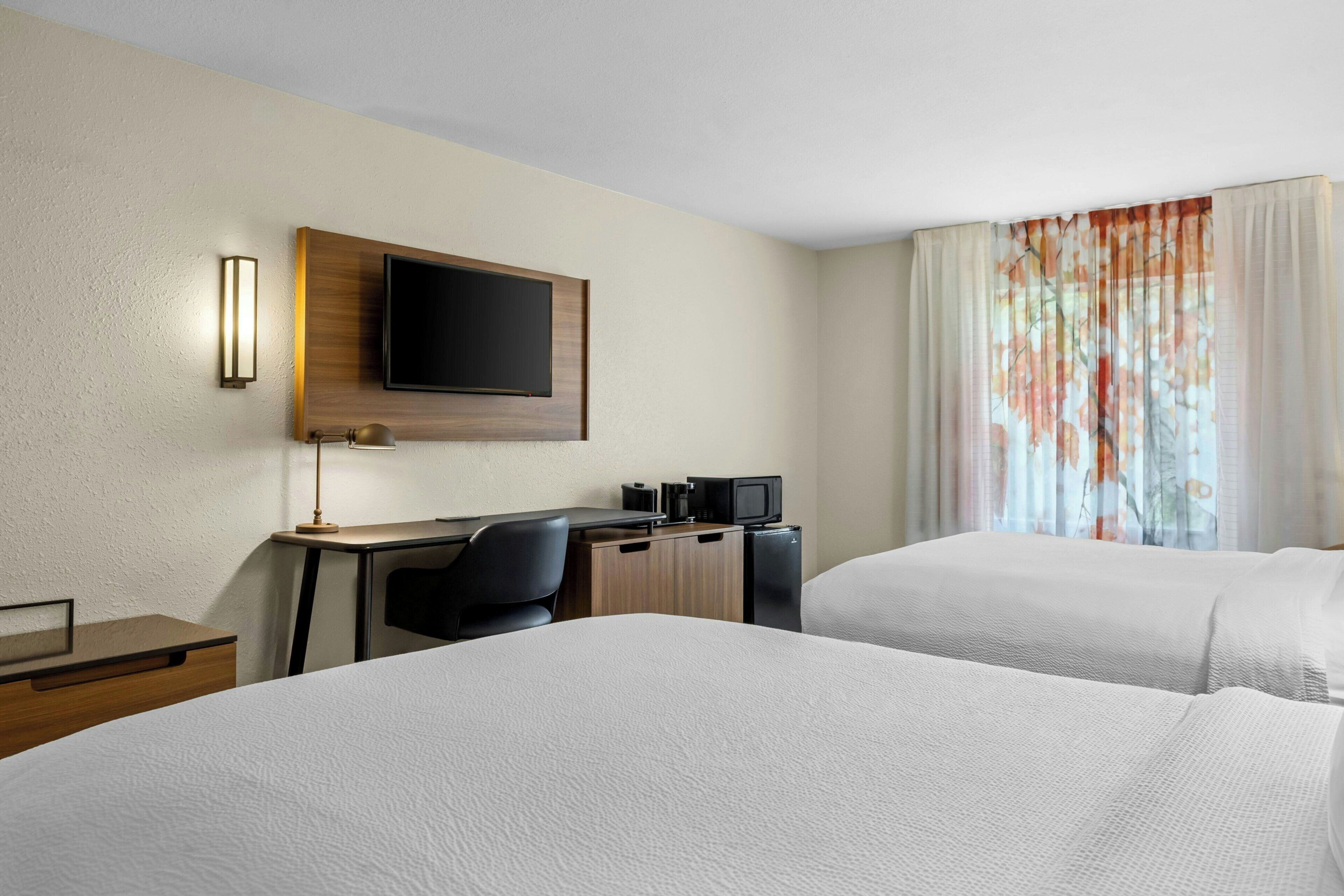 A hotel room with two beds, a desk with a TV, a desk lamp, and a small refrigerator.