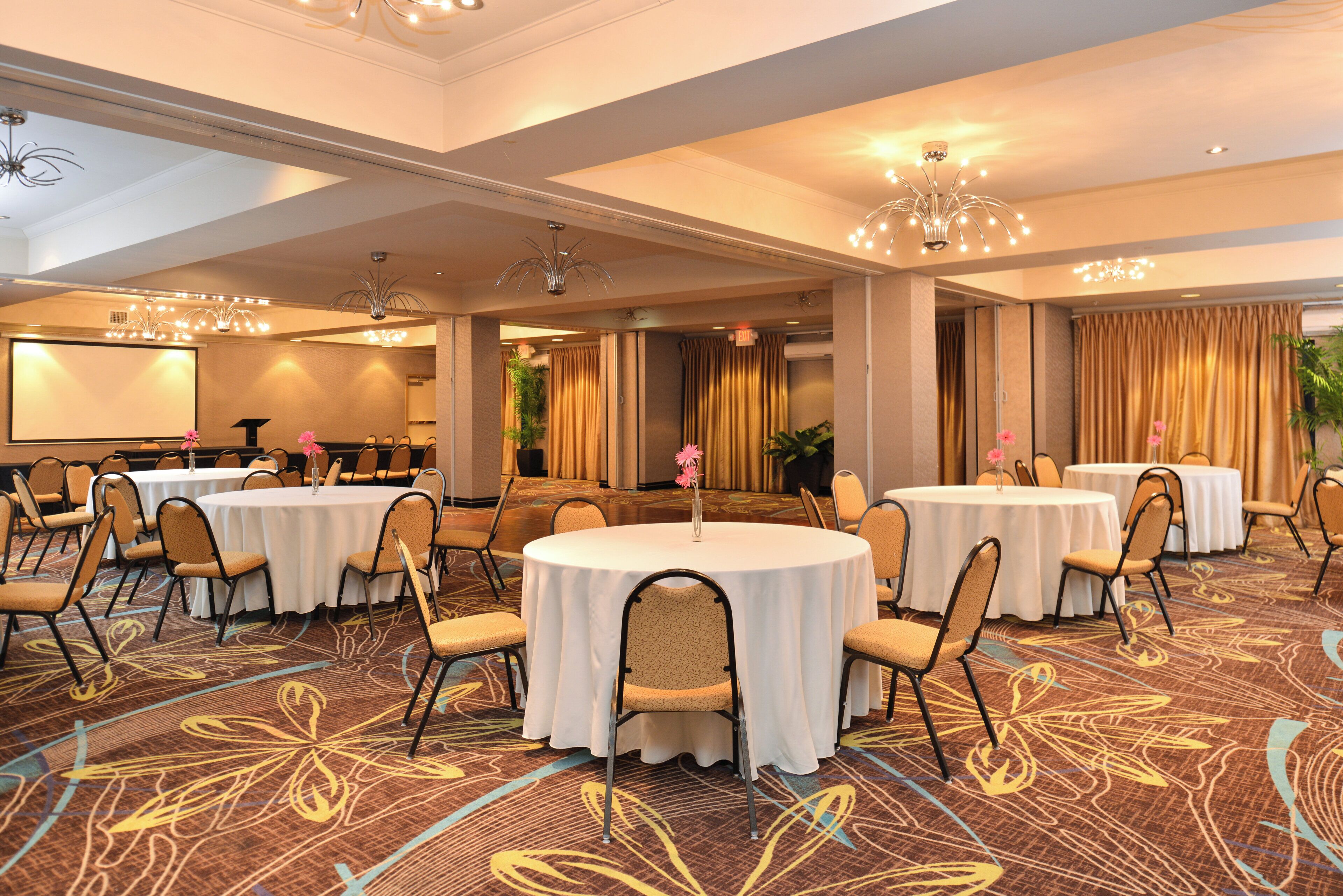 A hotel conference room with multiple round tables, chairs, and a projector screen.