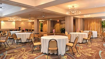 A hotel conference room with multiple round tables, chairs, and a projector screen.