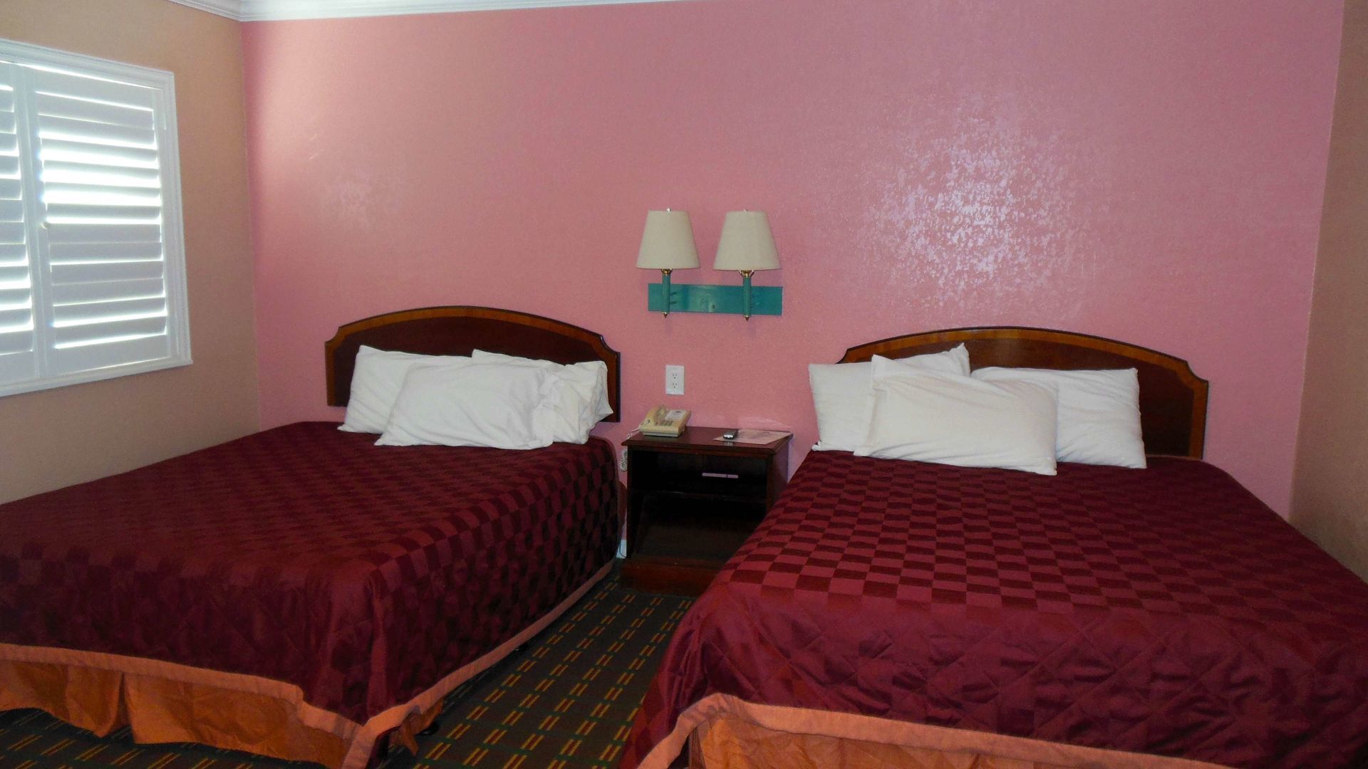A hotel room with two single beds, each with a wooden headboard and a checkered bedspread. There is a nightstand with a lamp between the beds, and a wall-mounted light fixture above the nightstand.