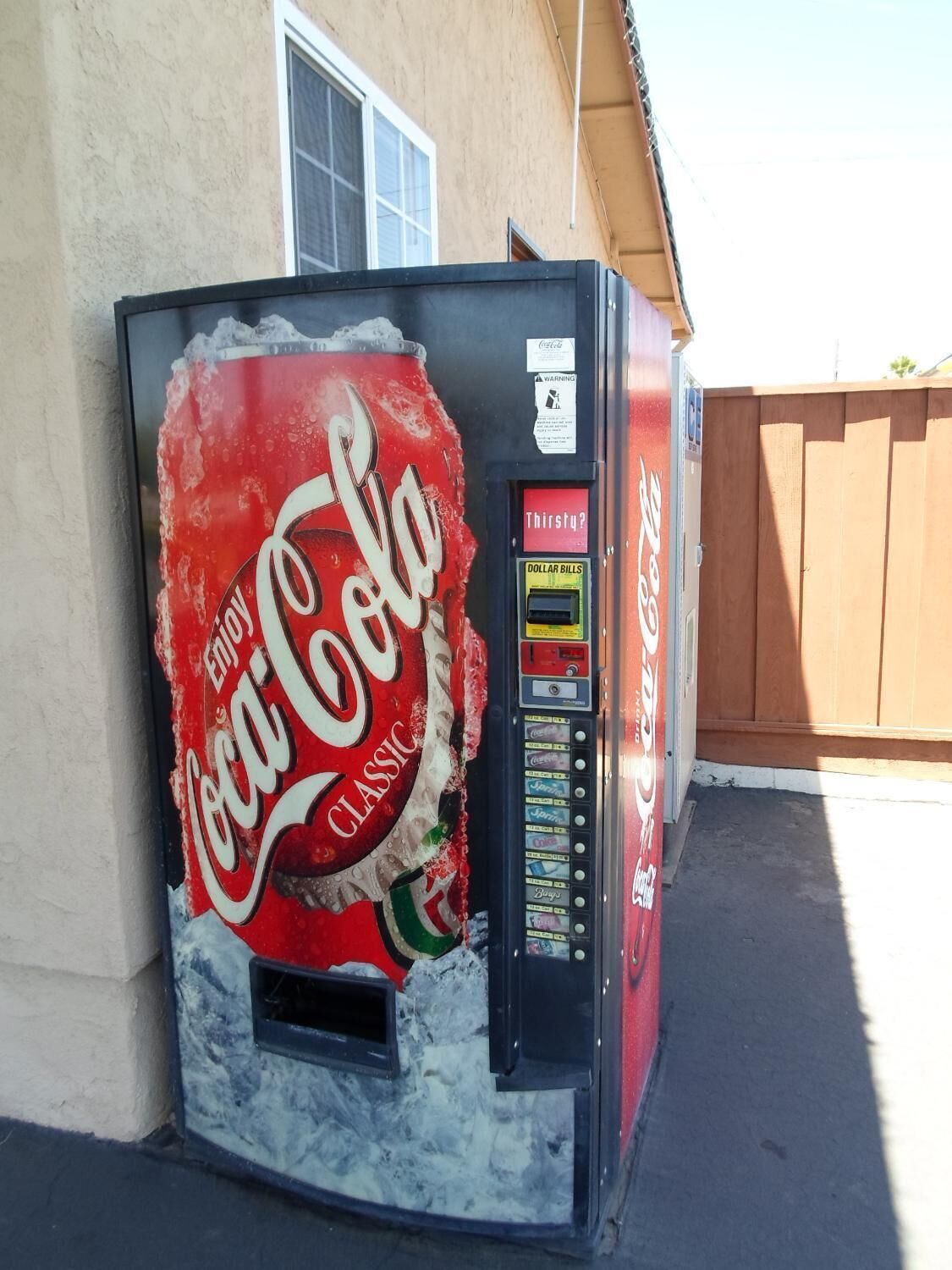 A Coca-Cola vending machine with a damaged front panel, a dollar bill dispenser, and a warning label.