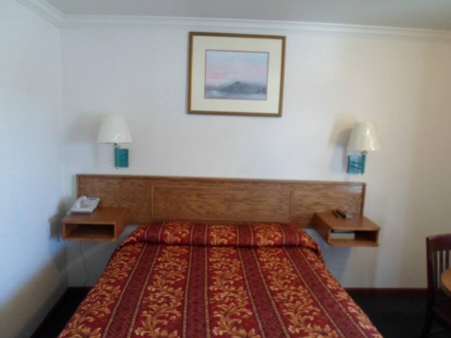 Un dormitorio con cabecera de madera, colcha estampada de color rojo, dos mesitas de noche con lámparas y un cuadro en la pared.