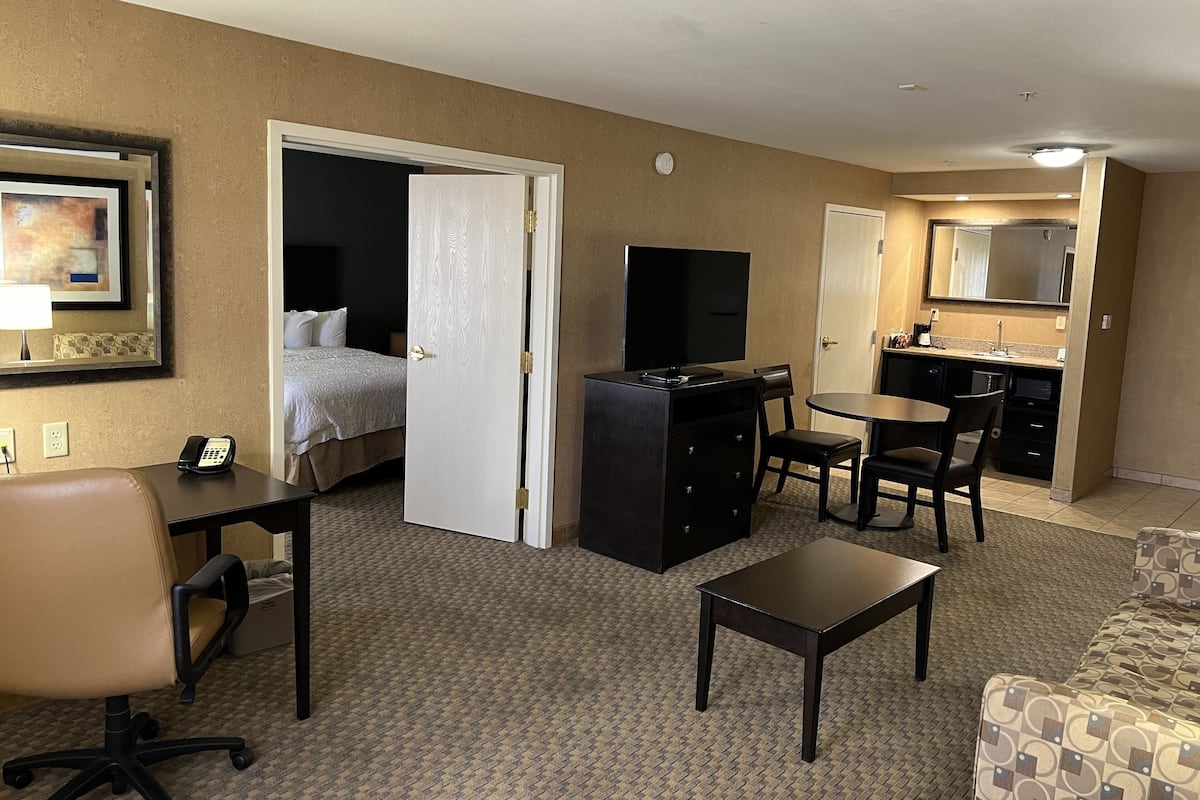 A hotel room with a bed, a desk with a chair, a small table, a sofa, a kitchenette, and a bathroom.
