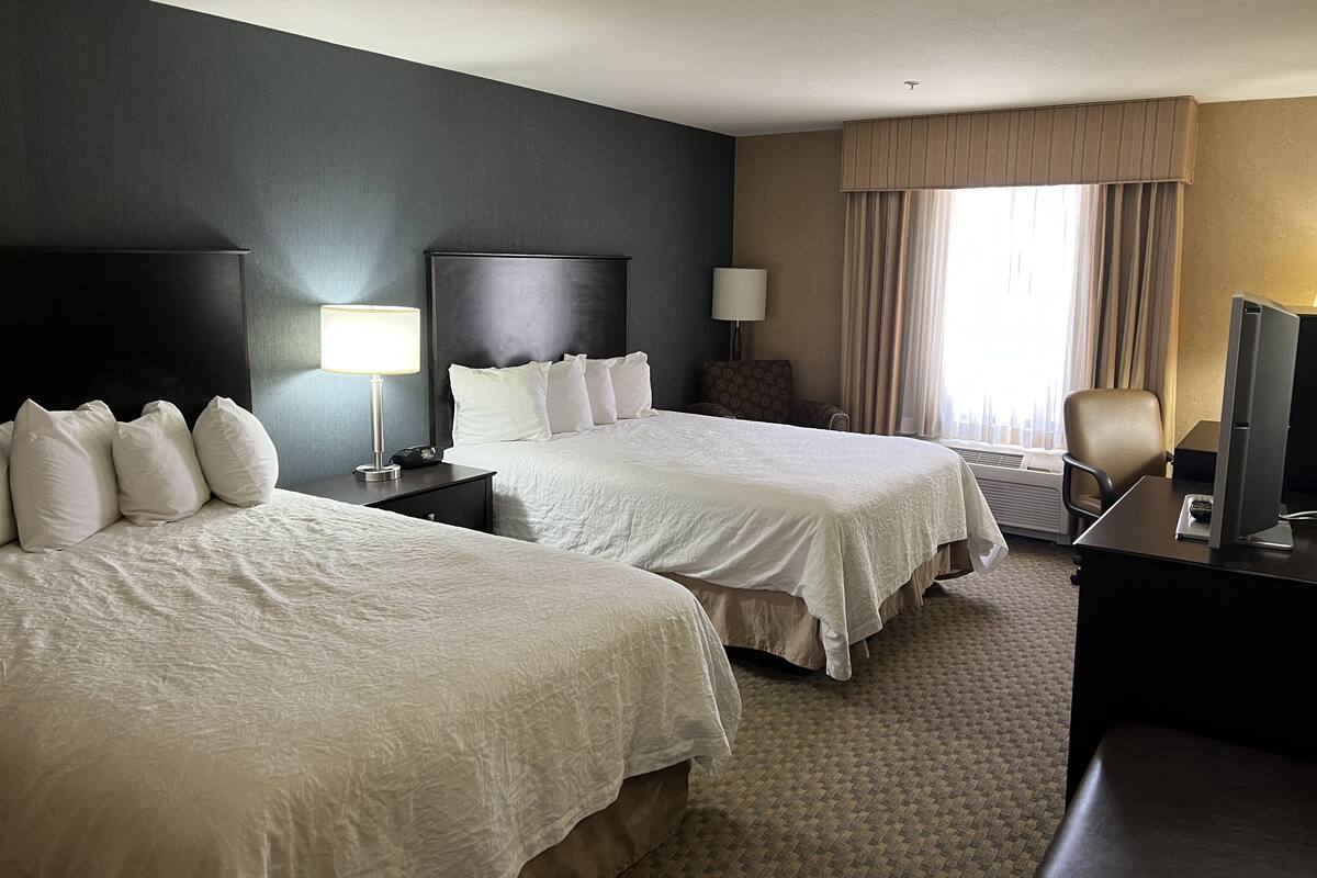 A hotel room with two beds, a desk with a computer, a chair, a lamp, and a window with curtains.