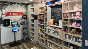 Zona de autopago con un letrero que dice 'Treats', un letrero que dice 'Bienvenido a Barstow' y estantes llenos de bocadillos y artículos de conveniencia.
