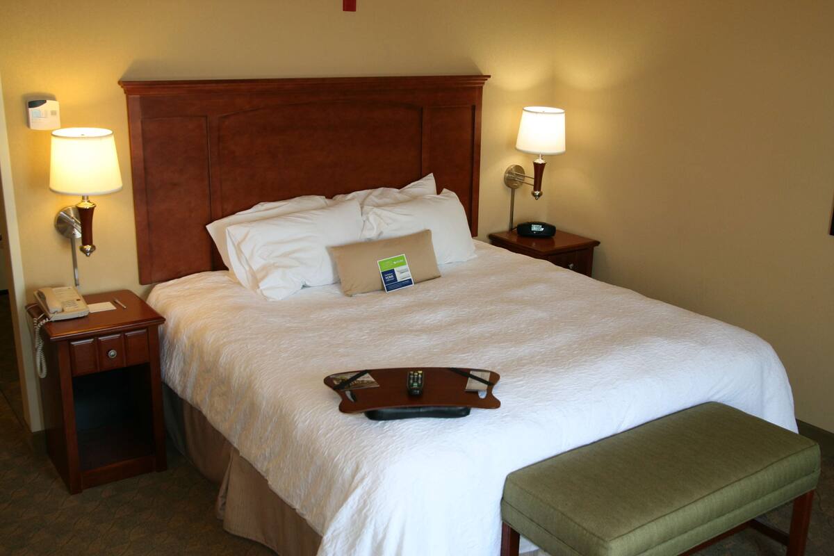 Una habitación de hotel con una cama grande, dos mesitas de noche, un banco verde y una bandeja con un control remoto.