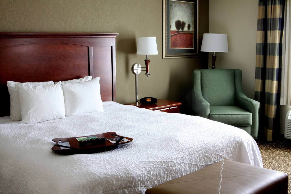 Una habitación de hotel con una cama grande, un sillón verde, una mesita de noche con una lámpara y una bandeja con un control remoto.