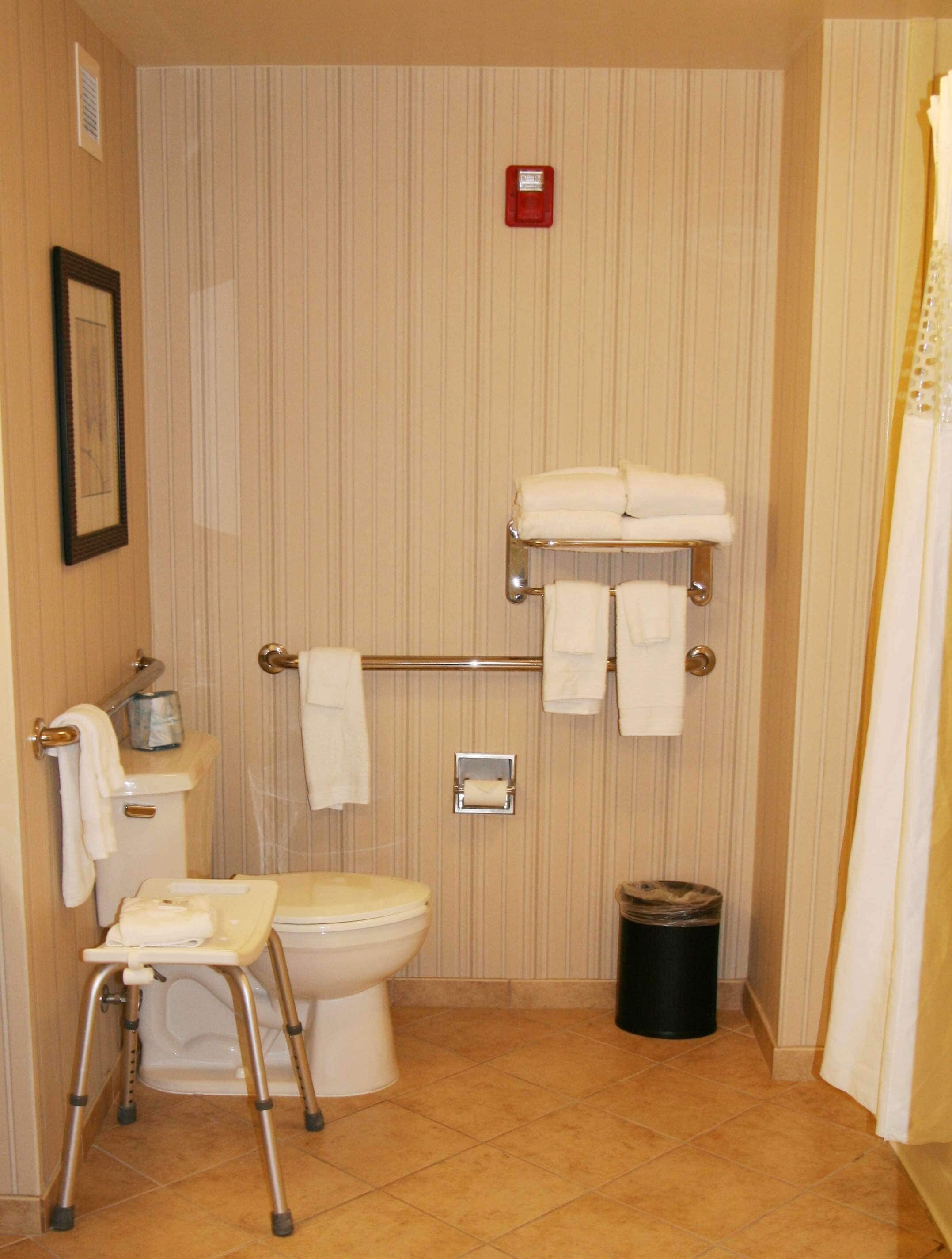 Une salle de bain équipée de toilettes, d’une barre d’appui, d’une poubelle et d’un fauteuil roulant.
