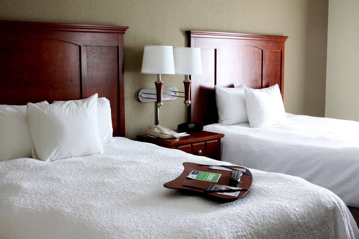 A hotel room with two beds, a bedside table with a phone and lamp, and a tray with a remote control.