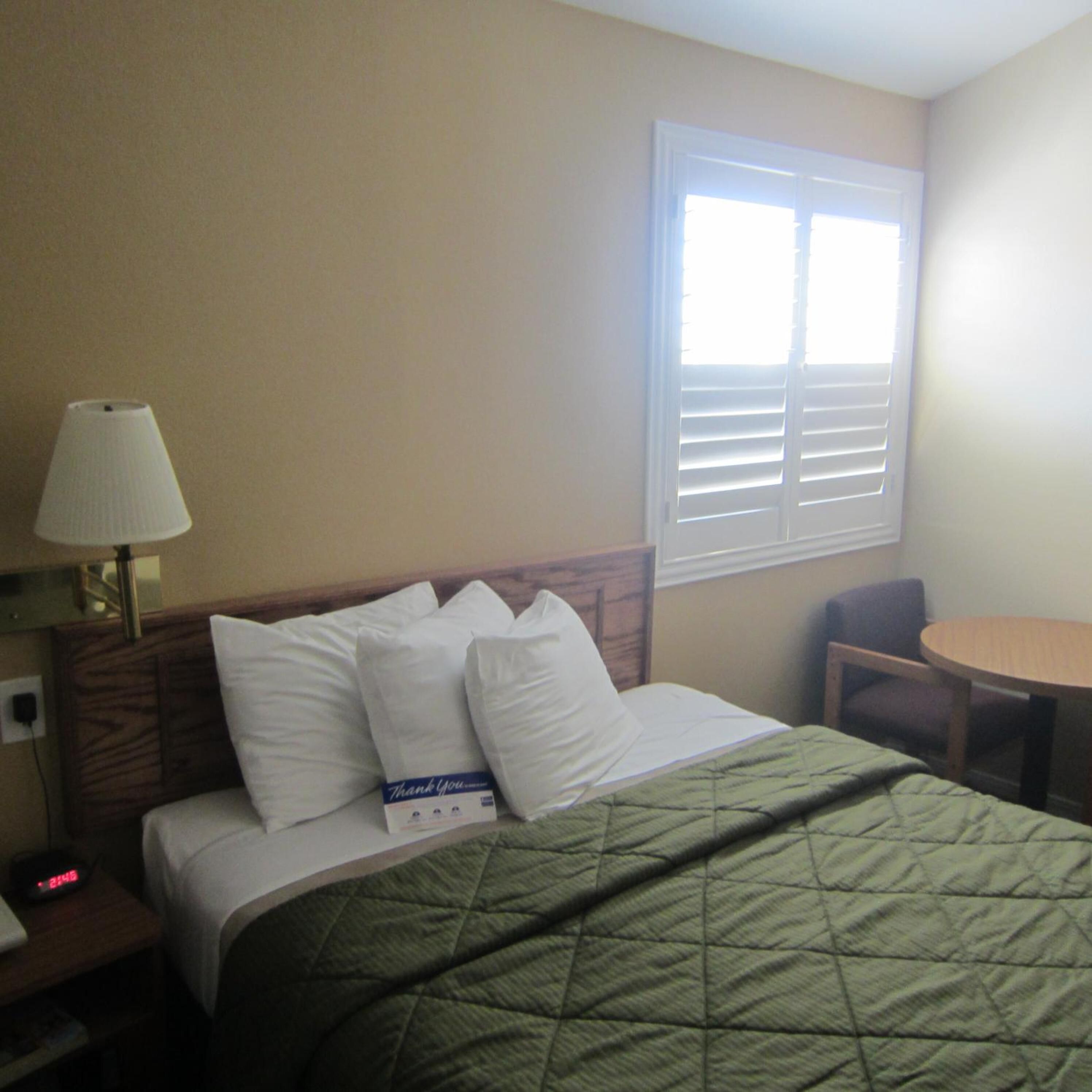 A hotel room with a bed, a bedside lamp, a window with blinds, a chair, and a small table.