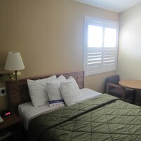 Una habitación de hotel con una cama, una lámpara de noche, una ventana con persianas, una silla y una mesita.
