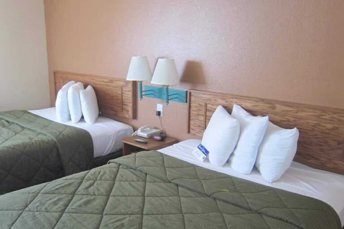 A hotel room with two beds, each with green quilts and white pillows. There is a nightstand with a lamp and a telephone.