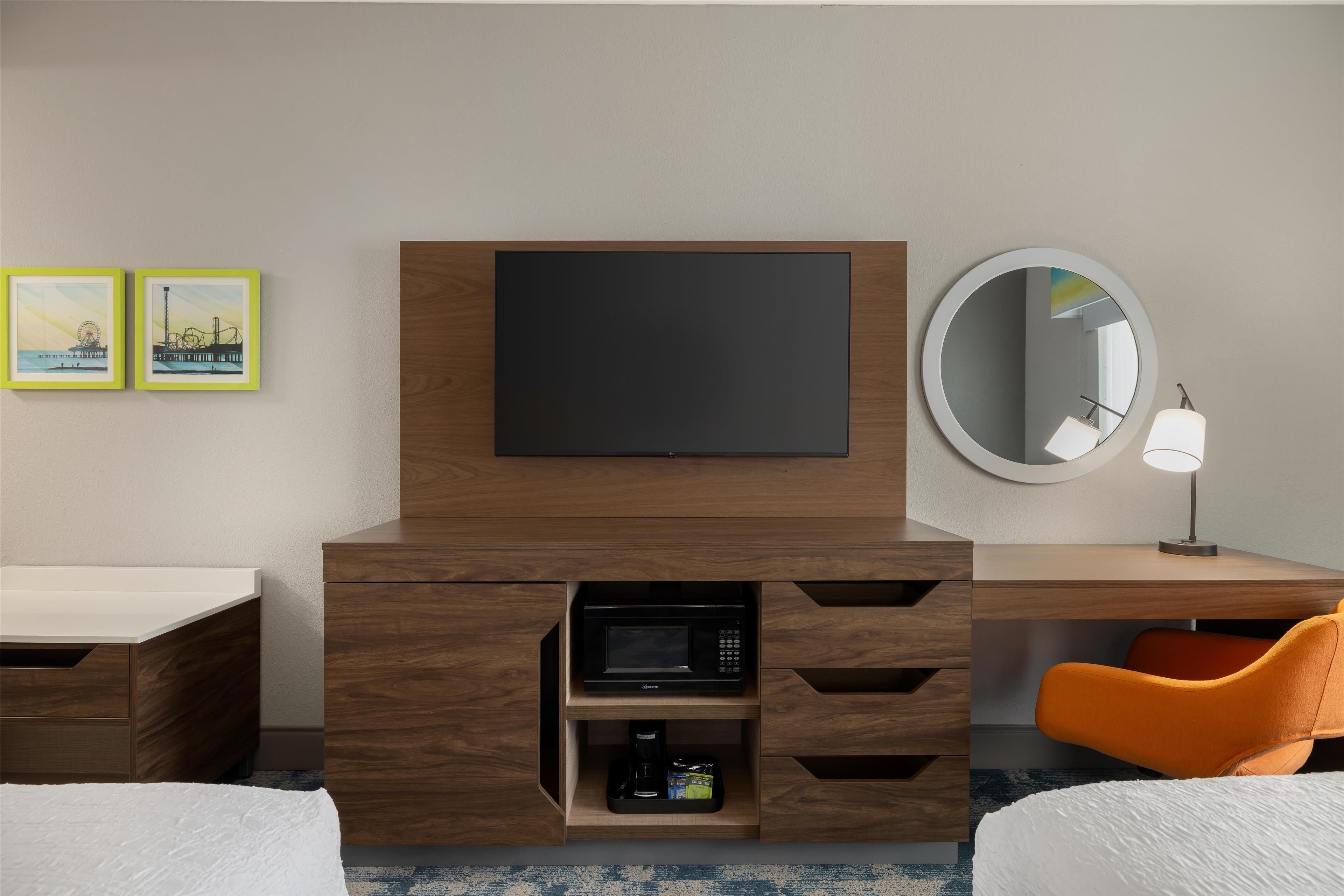 Habitación de hotel con un escritorio de madera, un televisor de pantalla plana, un microondas, una cafetera y una silla naranja.