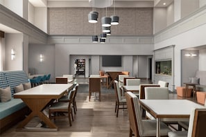 A spacious dining area with multiple tables and chairs, a high ceiling with hanging pendant lights, and a brick wall in the background. - Hampton Inn & Suites Galveston (Galveston)