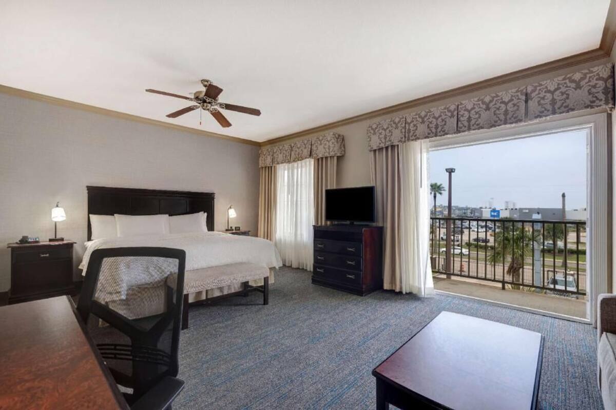 A hotel room with a bed, a television, a desk, a chair, a dresser, and a balcony with a city view.