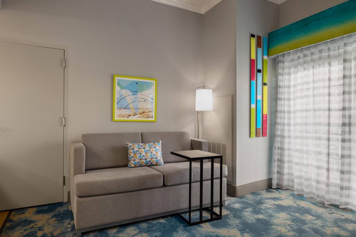 A modern hotel room with a grey sofa, a small table, a lamp, a colorful abstract painting, and a window with sheer curtains.