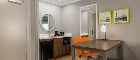 Una cocina moderna con microondas, pileta y pequeño refrigerador. Hay una mesa de madera con una silla naranja, un espejo redondo y dos cuadros en la pared.