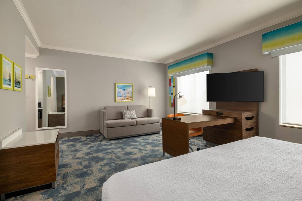 A hotel room with a bed, a desk, a sofa, a TV, and artwork on the walls.