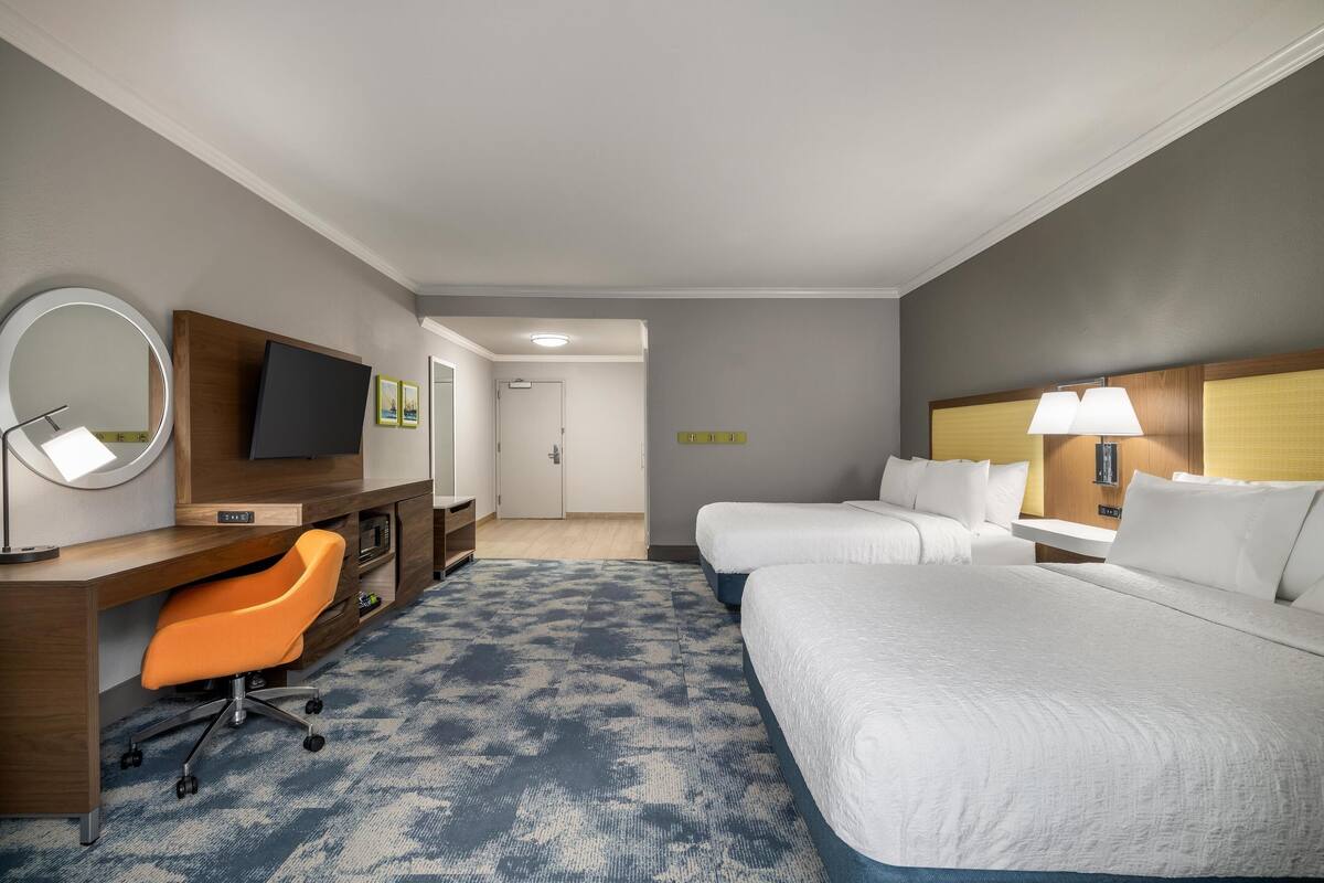 A hotel room with two beds, a desk with a chair, a TV, and a bathroom visible in the background.