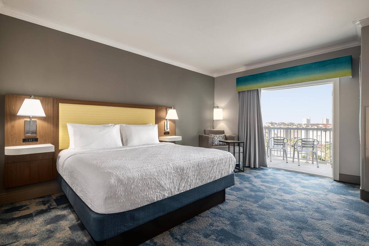 A hotel room with a large bed, two bedside tables with lamps, a small desk, and a balcony with a view of the city.