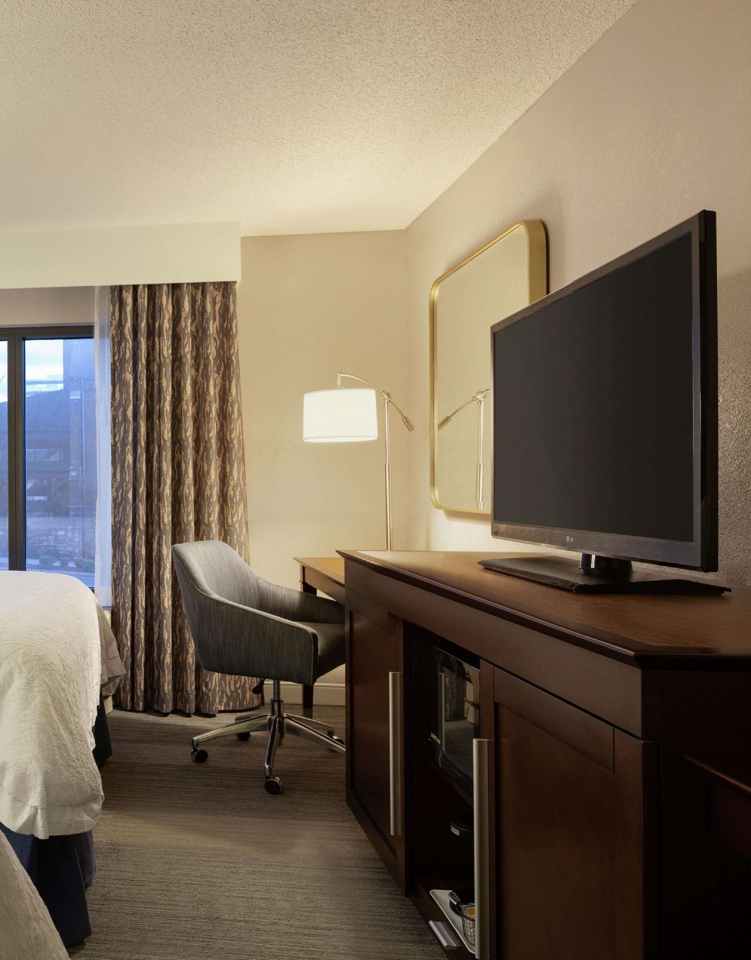 A hotel room with a bed, a desk with a television, a chair, and a large window with curtains.