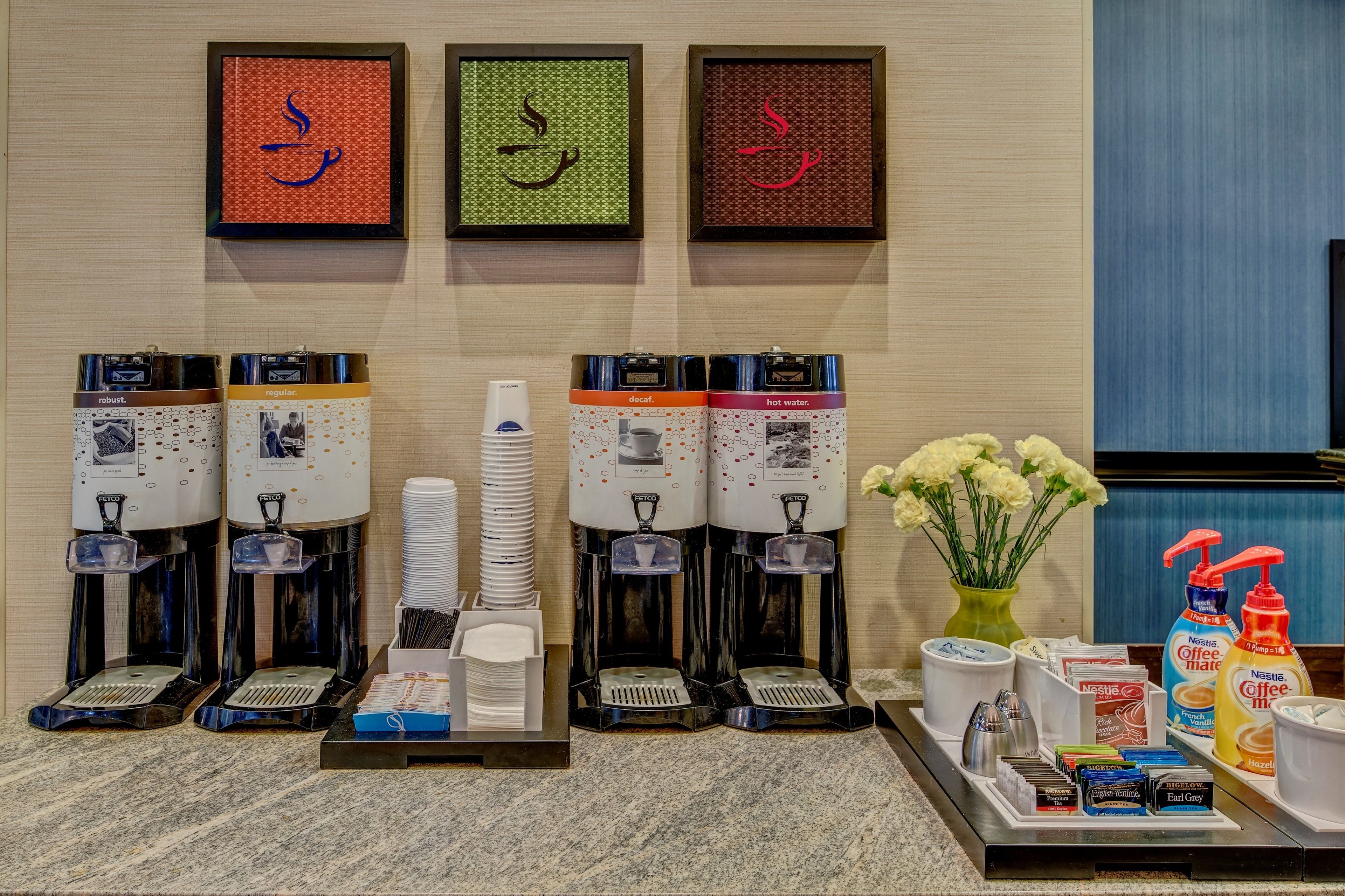 A hotel reception area with multiple beverage dispensers, a stack of white plates, and a tray with various items including a vase of flowers, a coffee maker, and a bottle of hand sanitizer.