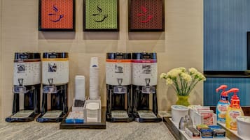 A hotel reception area with multiple beverage dispensers, a stack of white plates, and a tray with various items including a vase of flowers, a coffee maker, and a bottle of hand sanitizer.