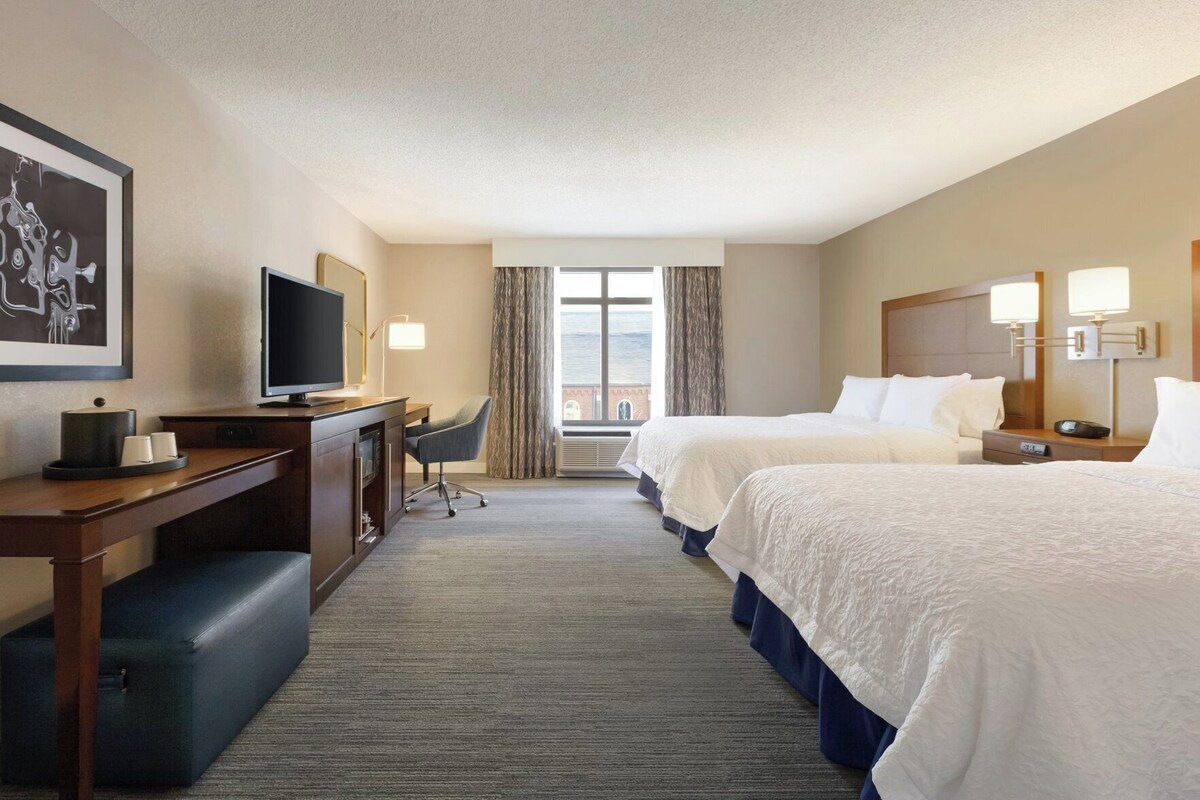 A hotel room with two beds, a desk with a TV, a chair, and a window with curtains.