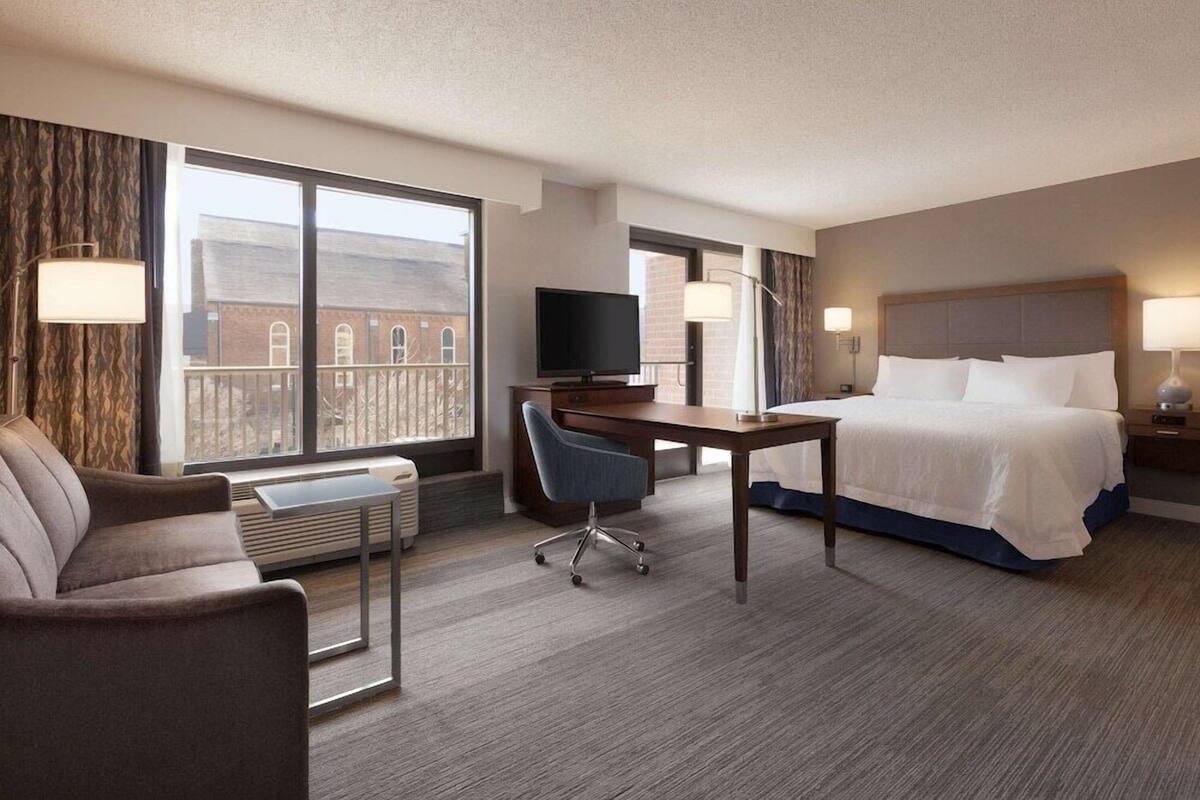 A hotel room with a bed, a desk with a chair, a TV, a lamp, and a view of a building through large windows.