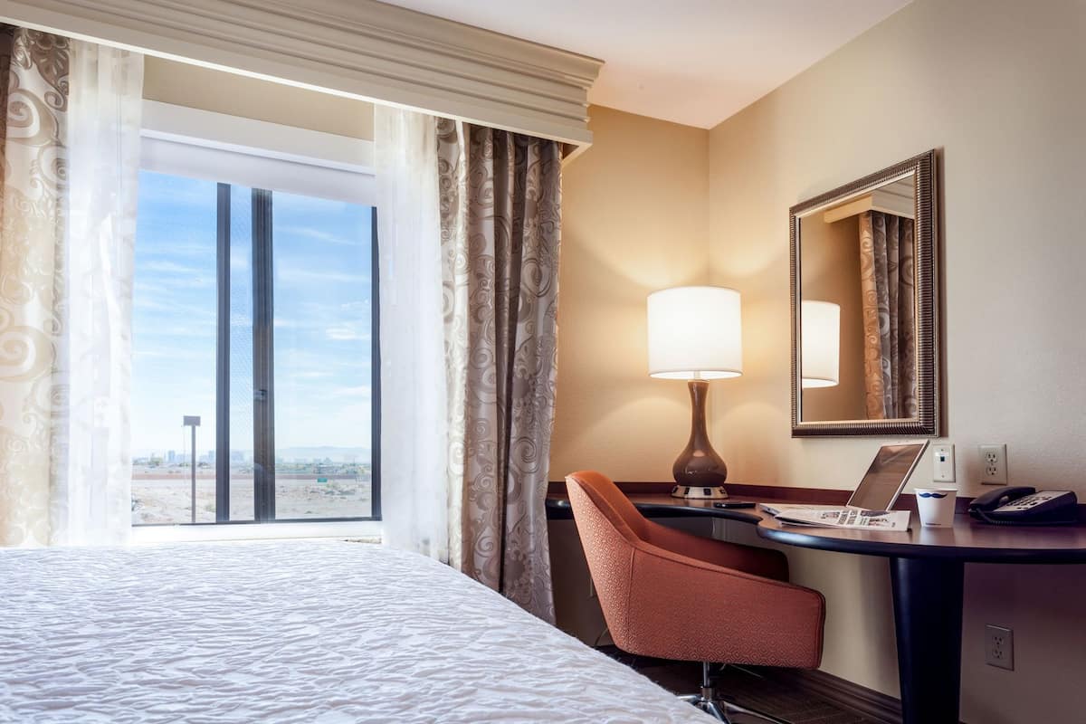 A hotel room with a bed, a desk with a lamp, a chair, a mirror, and a view of the desert through the window.