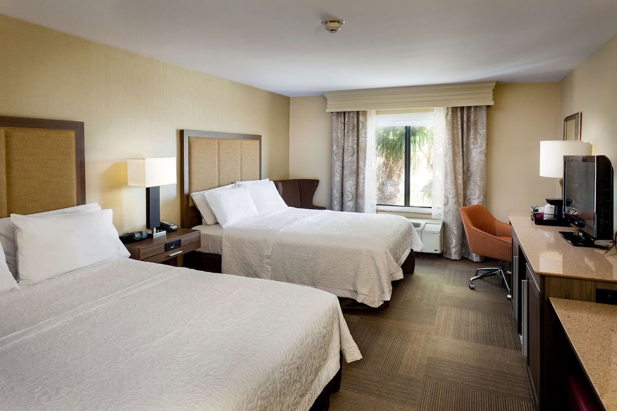 A hotel room with two beds, a desk with a computer, a chair, a window with curtains, and a television.