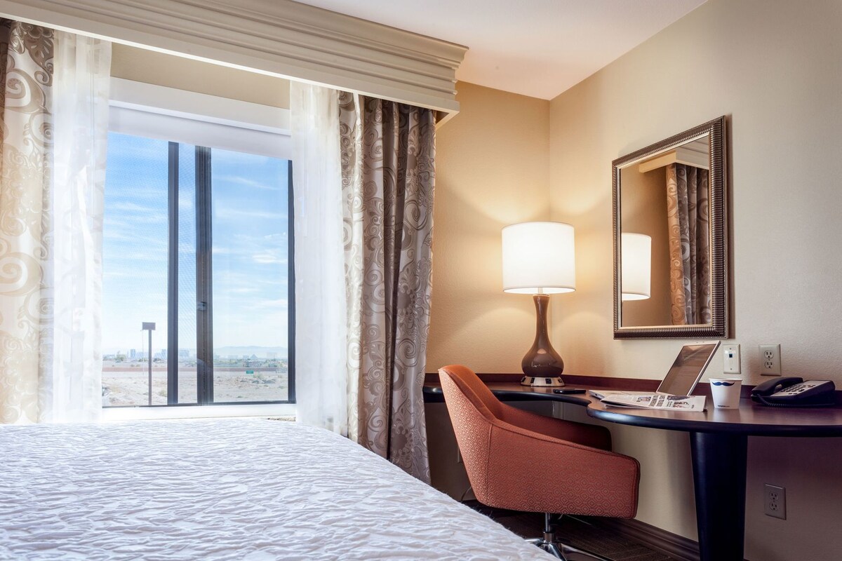 A hotel room with a bed, a desk with a lamp, a chair, a mirror, and a view of the desert through the window.