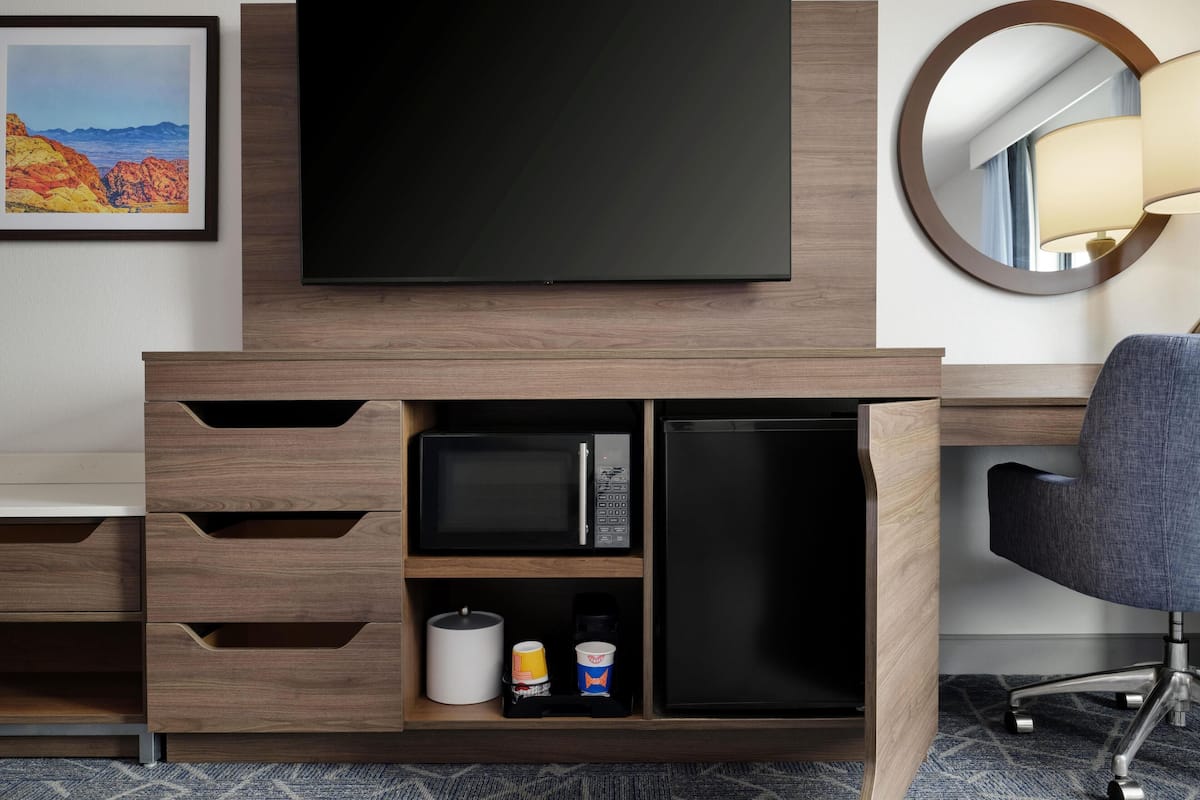 A hotel room with a flat-screen TV mounted on a wooden cabinet, a microwave, and a refrigerator drawer. There is a chair and a round mirror on the wall.