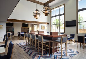 A modern dining area with a long wooden table and bar stools, large windows, and a patterned rug. - Hampton Inn & Suites Las Vegas-Red Rock/Summerlin (Las Vegas)