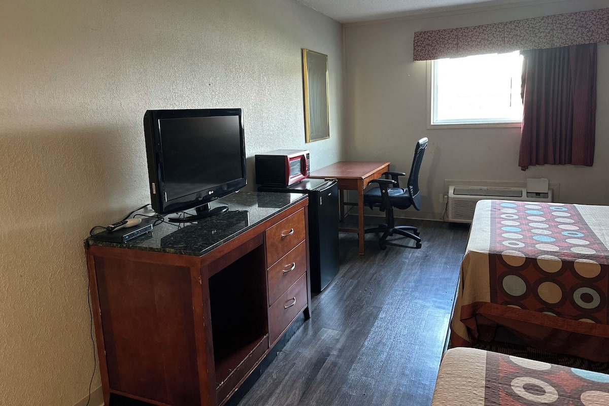 A room with a wooden desk, a black LG monitor, a red and black speaker, a black office chair, a patterned bedspread, and a window with curtains.