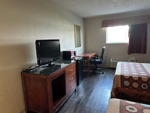 A room with a wooden desk, a black LG monitor, a red and black speaker, a black office chair, a patterned bedspread, and a window with curtains. - Airport Inn Flint (Flint)
