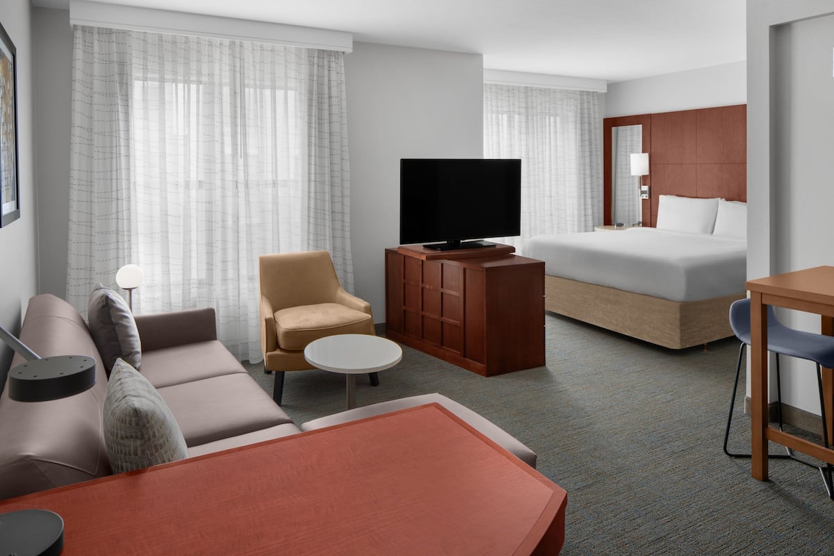 Habitación de hotel con sofá, sillón, mesita redonda, televisor sobre un mueble de madera, cama con cabecero de madera y escritorio con silla.