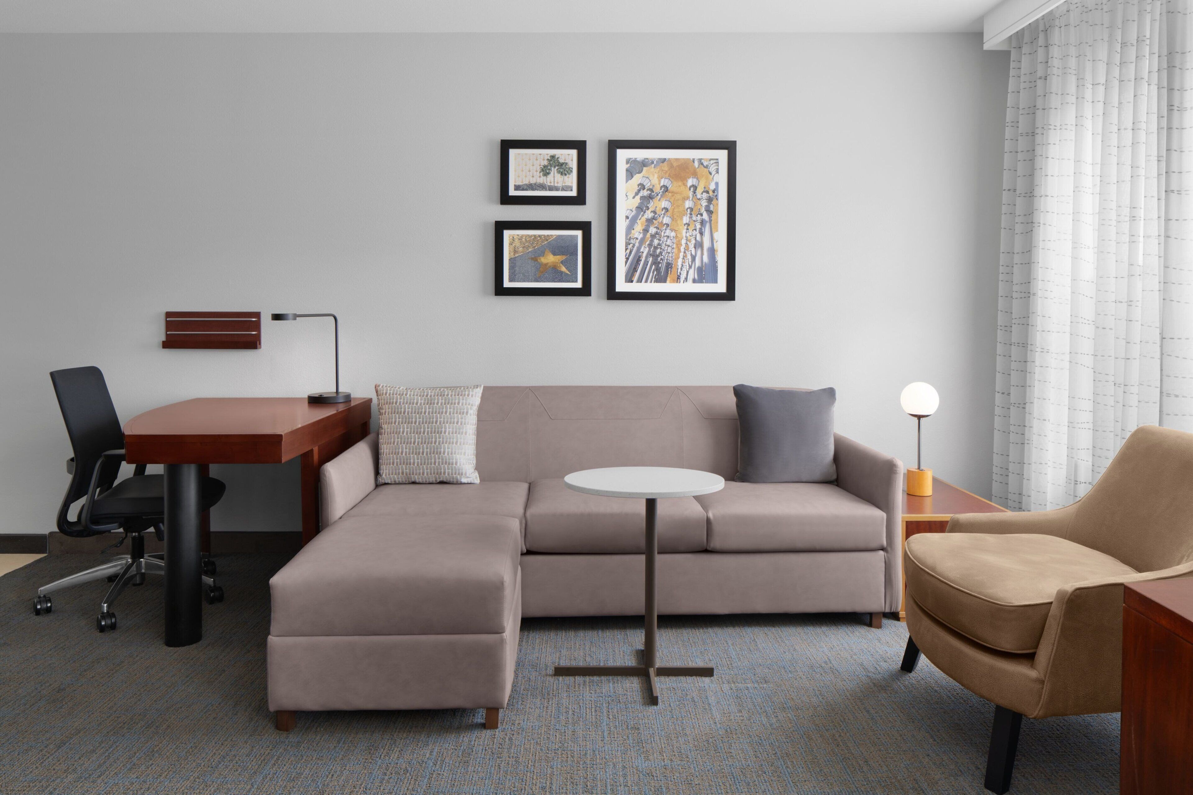 Ein modernes Hotelzimmer mit einer Couch, einem kleinen runden Tisch, einem Schreibtisch und einem Sessel. An der Wand hängen drei gerahmte Kunstwerke.