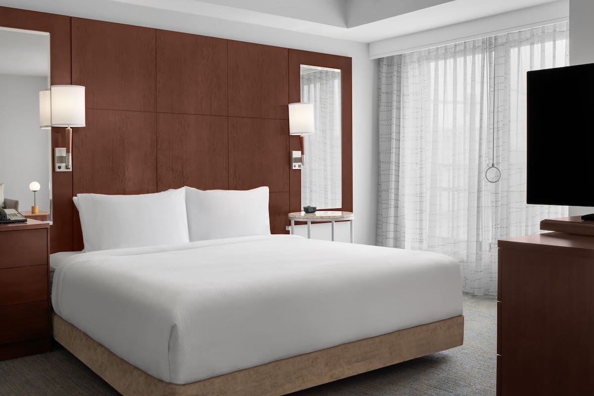 Habitación de hotel con una cama grande, cabecero de madera, lámparas de noche, una mesita, un televisor y un ventanal con cortinas traslúcidas.