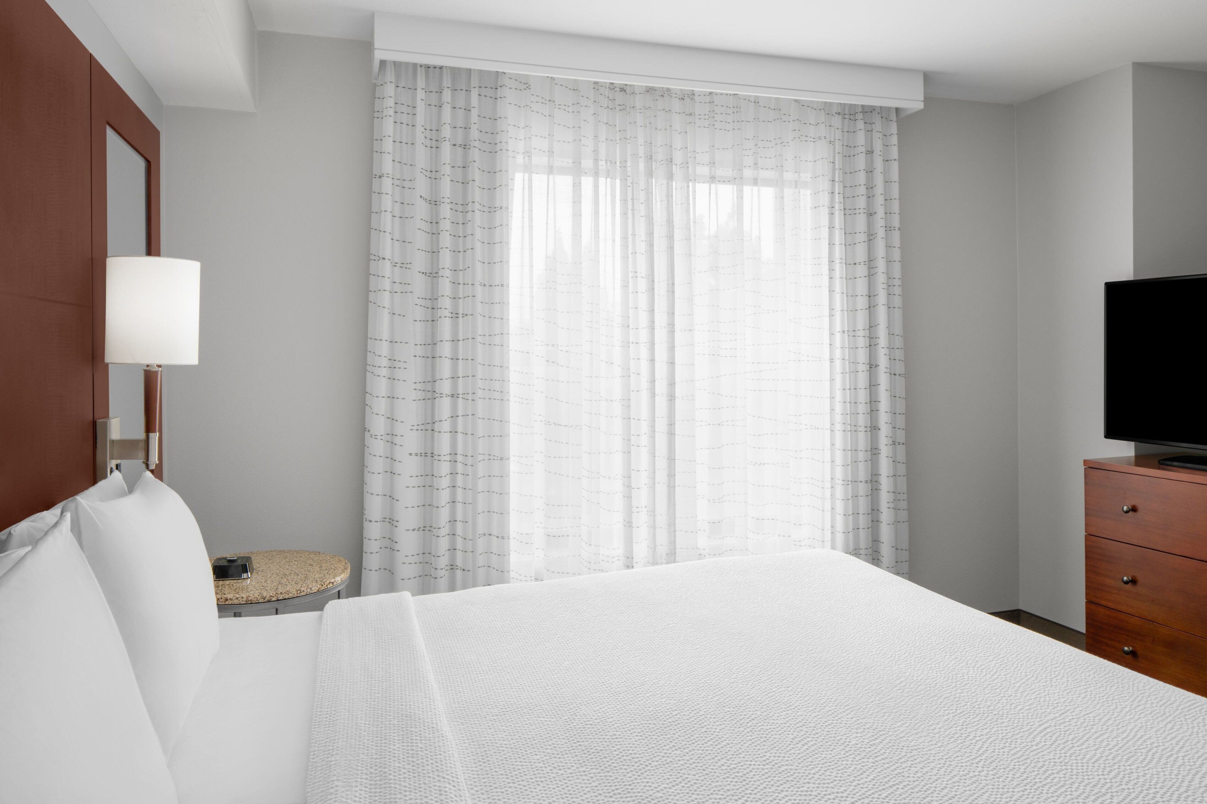 Habitación de hotel con cama, televisor sobre una cómoda de madera, una mesita redonda y un gran ventanal con cortinas traslúcidas.