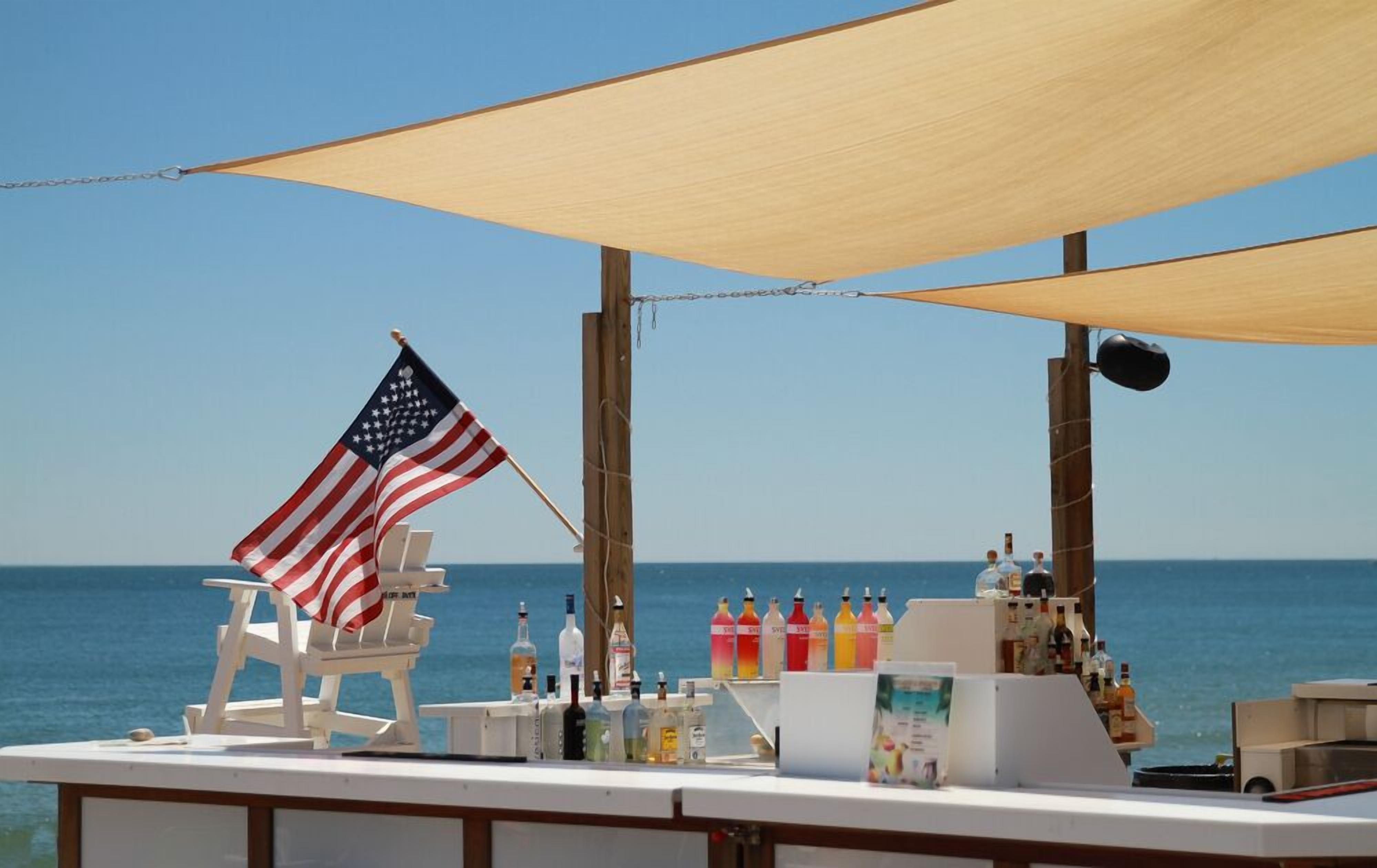 A beach bar with an American flag, a white chair, and various bottles on the counter.