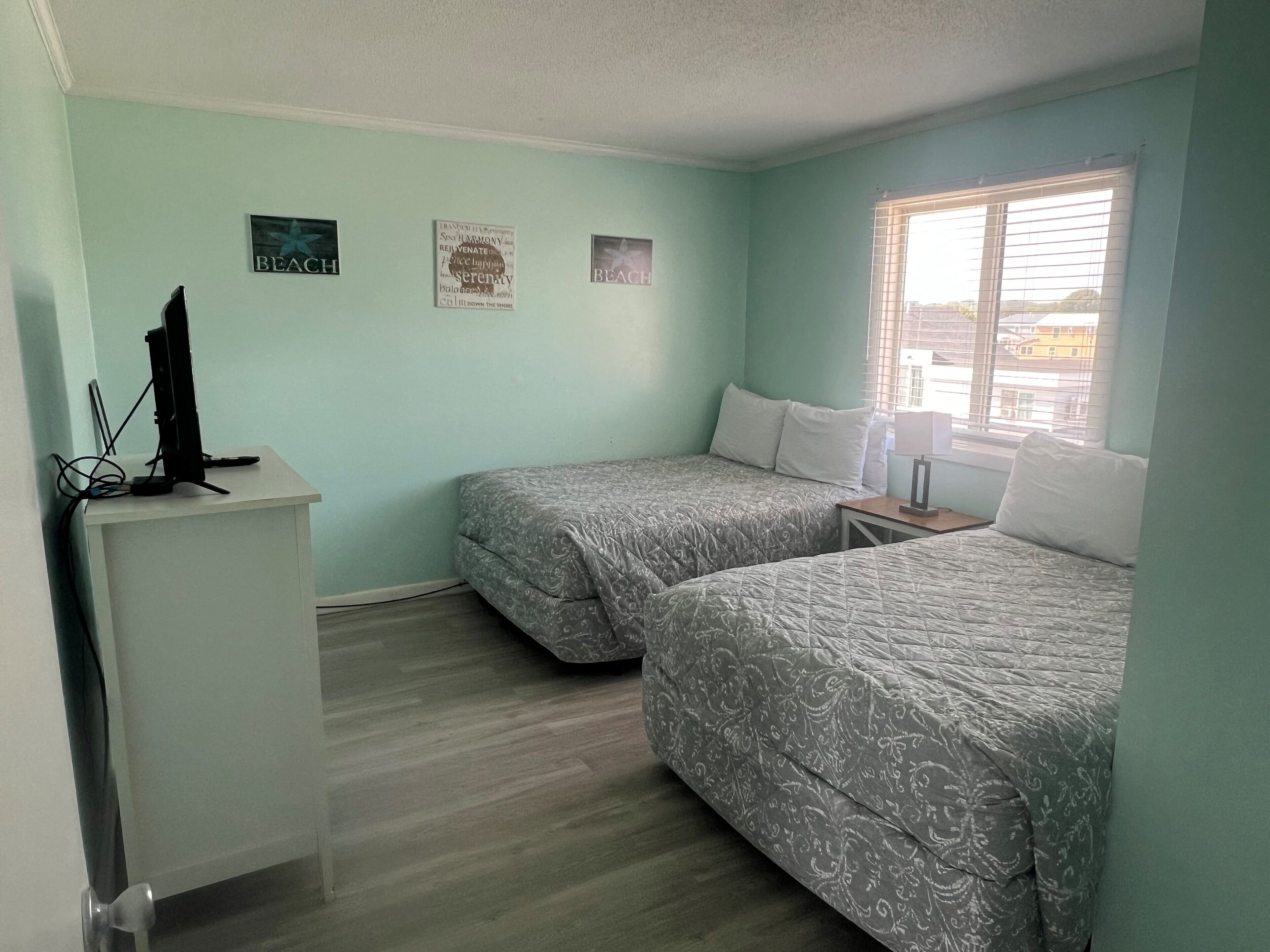 Ein Zimmer mit zwei Betten, einem Schreibtisch und einem Computer. Zwei Wandbilder und ein Fenster mit Jalousien sind vorhanden.