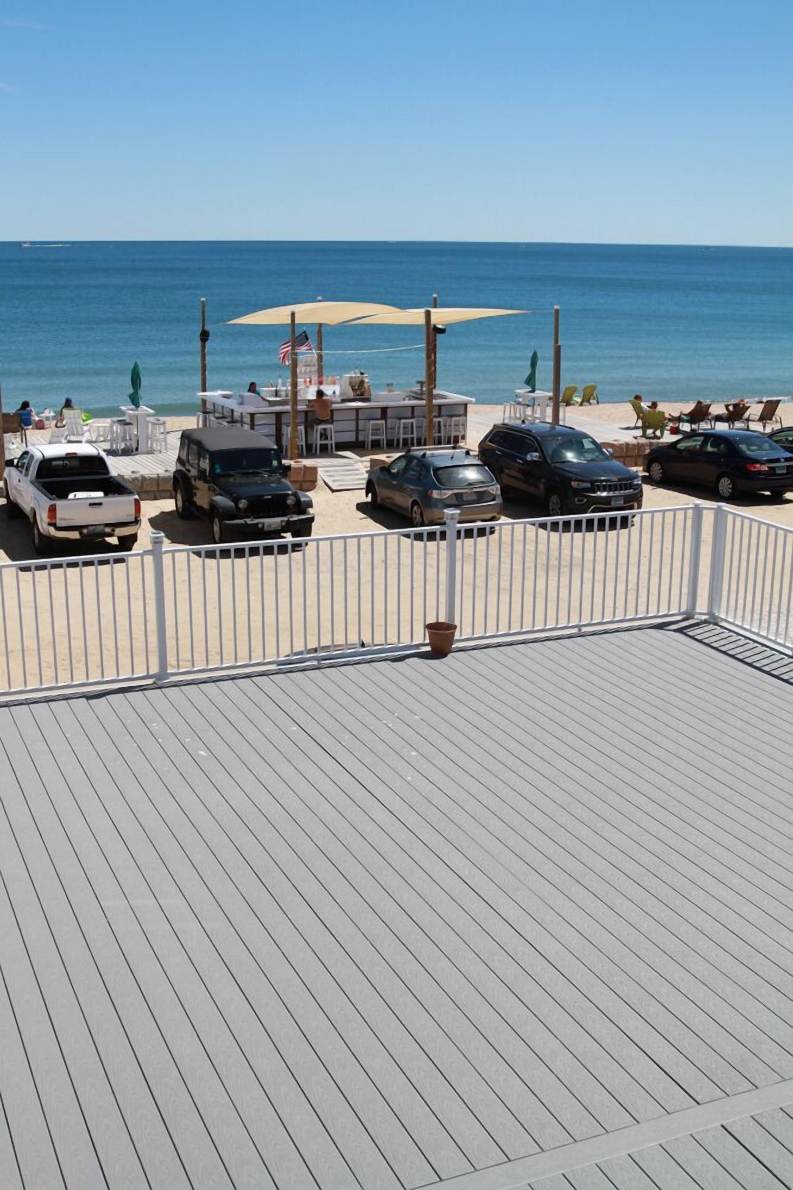 Terraza frente a la playa con vista al océano, varios vehículos estacionados y zonas de estar al aire libre.